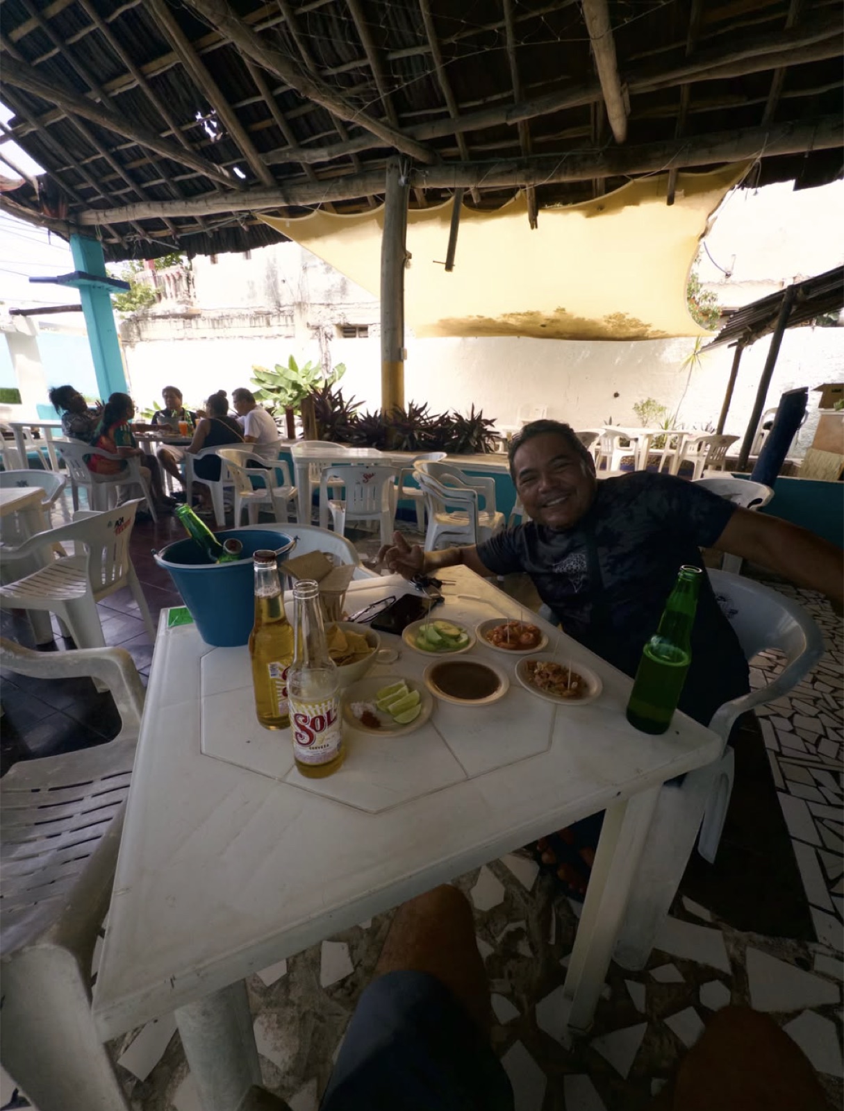 Miguel grinning at a white plastic table at William’s in Cozumel, under a palapa roof, with two Sol beers, a bucket of ice, lime wedges, and small plates of snacks in front of him